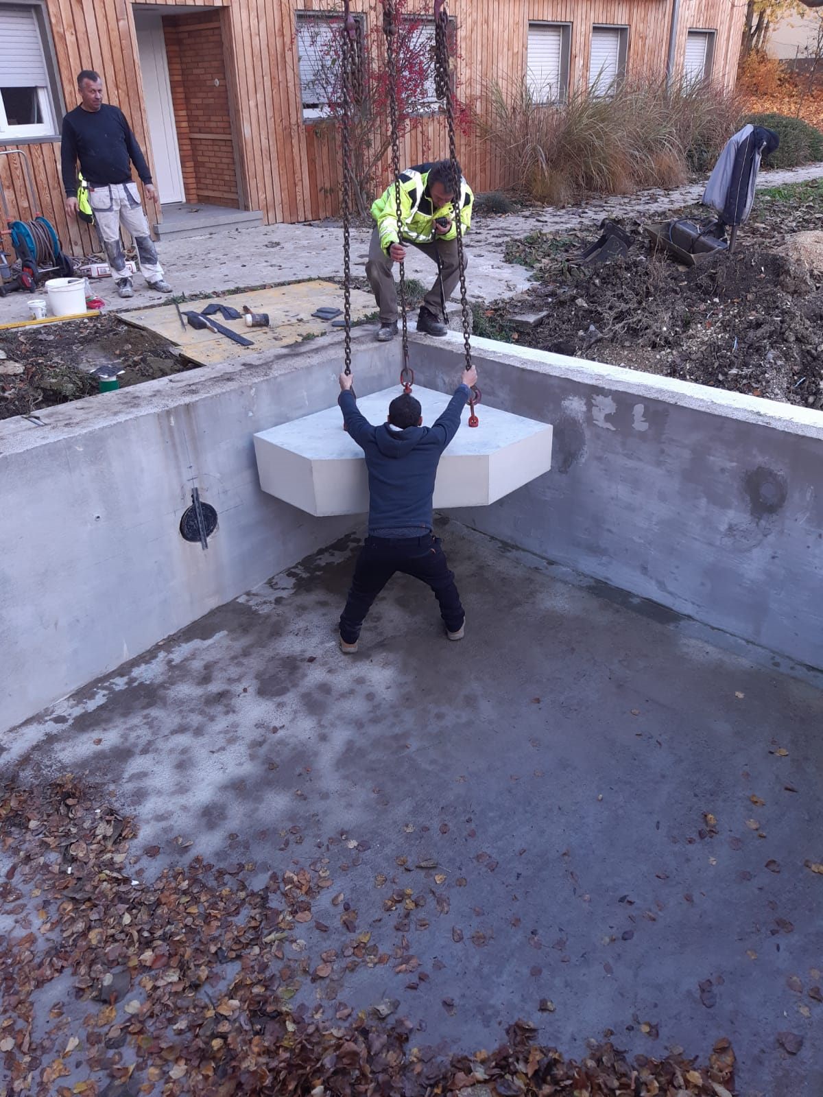 Drei Mitarbeiter setzen ein Betonfertigteil mit dem Kran in den Rohbau eines Pools ein